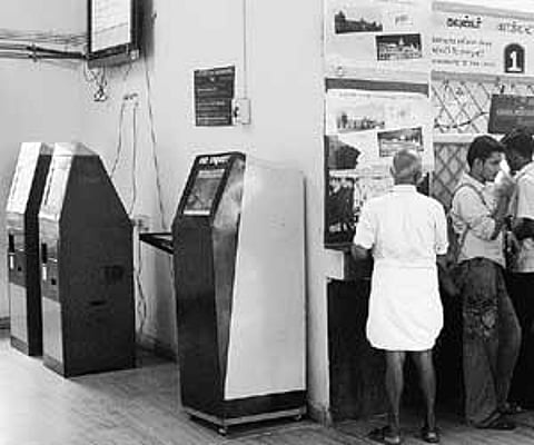 People standing in a queue to collect tickets at a counter in Egmore suburban station, while the automatic vending machines adjacent to the counter re