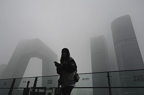 A woman uses her phone along a glass wall with the China Central Television (CCTV) headquarters in the background on a polluted day in Beijing (Photo | AFP)