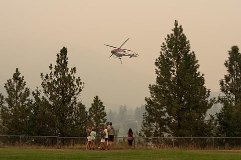 People gather to a lookout as a helicopter gathers water from Shannon Lake to fight the McDougall Creek wildfires in West Kelowna, British Columbia on August 19, 2023. (Photo | AFP)