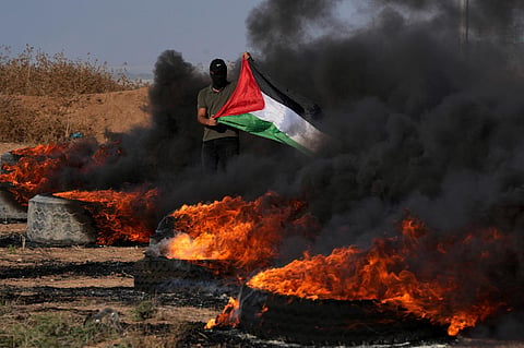 A masked Palestinian protester waves his national flag next to burning tires during clashes with Israeli security forces along the frontier with Israel. (Photo | AP)
