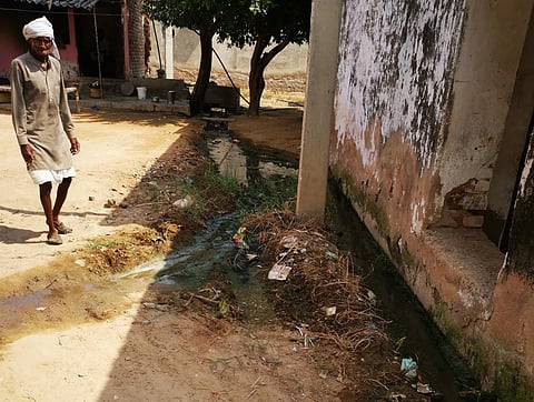 A man stands in front of a house in rural India as grey water flows next to it in the open, creating waterlogging.