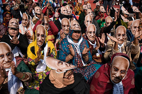 A large group of women are sitting together, wearing masks of Mohandas Karamchand Gandhi's face, participating in a peaceful demonstration.