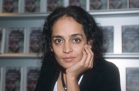 Arundhati Roy rests her chin on her hand and looks directly at the camera. She is seated indoors in front of shelves filled with multiple copies of 'The God of Small Things', whose covers are blurred in the background.