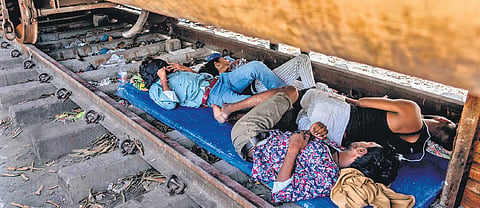 Roadside vendors rest in the shade beneath a stationary train coach on a hot summer day in Mumbai