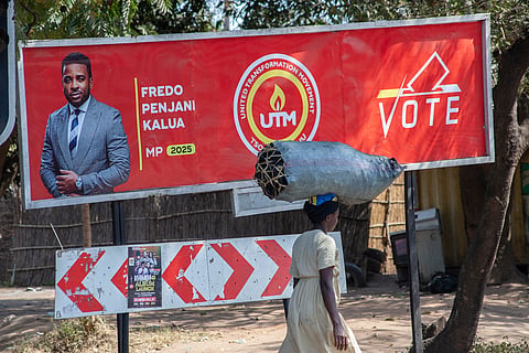 A woman carrying a bag of charcoal walks past a billboard calling on constituents to vote for aspiring UTM Parliamentary candidate Fredo Penjani Kalua, also known as Fredokiss, in Blantyre on August 9, 2025.