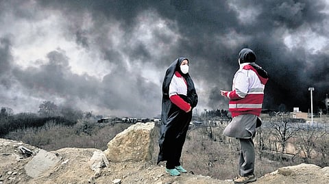 Two women from the Iranian Red Crescent Society stand as a thick plume of smoke from a US-Israeli strike on an oil storage facility rises in Tehran, Iran.