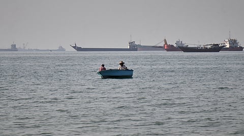 Two men sit in a small boat on the water as a mix of bulk carriers, cargo ships, and service vessels line the horizon in the Strait of Hormuz off Bandar Abbas, Iran, Monday, April 27, 2026.