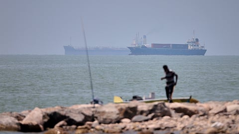 A man stands beside a fishing pole along the shore as cargo ships and commercial vessels are seen in the Strait of Hormuz off Bandar Abbas, Iran, Wednesday, June 17, 2026.