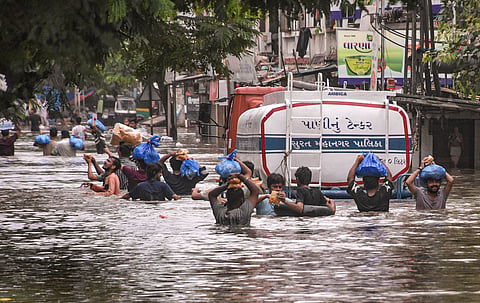 People carry packets of food items on their heads as they wade through a waterlogged road after heavy rain, in Surat, Wednesday, July 8, 2026.