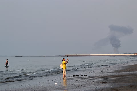 A woman stands at the water's edge along the Strait of Hormuz as a plume of smoke rises in the background following an explosion, off Bandar Abbas, Iran, Monday, July 13, 2026.