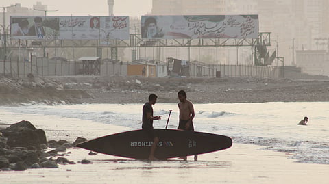 Two men stand with a surfboard along the shore in the Strait of Hormuz off Bandar Abbas, Iran, as billboards depicting Iran's late Supreme Leader Ayatollah Ali Khamenei rise in the background, Monday, July 27, 2026.