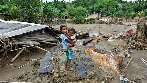 A little girl holds her younger sister, who stands on debris and thick mud, where devastating floods destroyed homes and livelihoods at Nepali Khuti in Sivasagar, Assam.