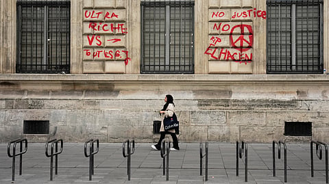 A woman walks past a building with graffiti that reads "The ultra-rich against everyone" and "No justice, no peace. RIP El Hacen" after the weekend protests over the cost of living turned into rioting and looting across France, in Paris, Wednesday, Oct. 21, 2026.