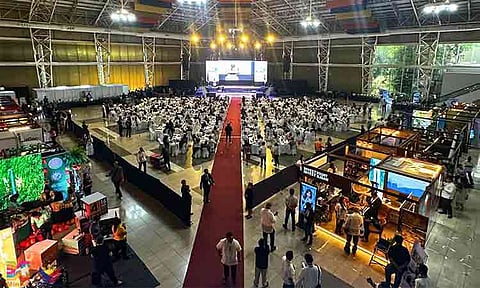 Delegates gather for a large-scale business convention inside the Limketkai Atrium in Cagayan de Oro City, Misamis Oriental, Philippines. The high-angle view captures the expansive indoor venue arranged with formal banquet tables, a central red carpet aisle leading to a main stage, and commercial exhibition booths promoting local economic initiatives.