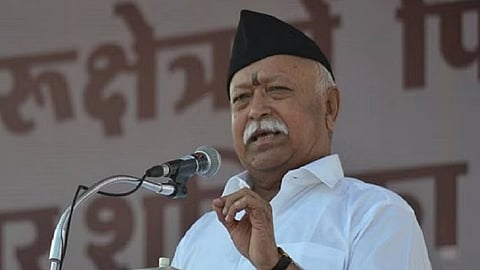RSS chief Mohan Bhagwat in a white shirt and black cap, speaking at a mic. Banners can be seen behind him.