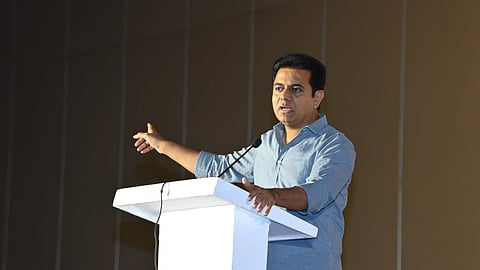 BRS leader KTR in a grey shirt standing at a podium speaking into a mic. The background is a brown wall with thing grey stripes. His right hand is on the podium, his left hand is pointing to his left side.