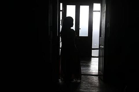 A silhouette of a young girl with long hair stands in a darkened doorway, facing away from the camera toward a brightly lit, misty background visible through windowed doors. The scene is high-contrast, with the foreground in deep shadow and the light source creating a glowing outline around her figure.