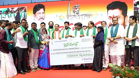 A group of people on a stage hold a large ceremonial check, with Telangana CM Revanth Reddy at the centre. The backdrop displays an illustration of a farmer and photos of CM Revanth and Dy CM Bhatti Vikramarka. People are dressed in formal and traditional attire, and there are floral decorations on stage.