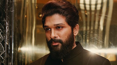 Allu Arjun in a black suit staring intensely to his left, standing against a shiny backdrop