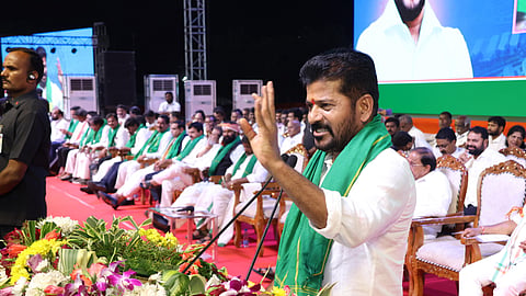 Telangana CM Revanth Reddy in a white shirt and green scarf speaks passionately at a podium adorned with flowers. People in white clothing are seated behind him on a stage, with a large screen displaying an image in the background.