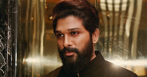 Allu Arjun in a black suit staring intensely to his left, standing against a shiny backdrop