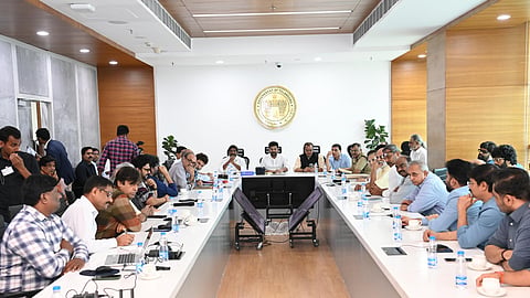 A group of men are seated around a long conference table in a modern meeting room. Telangana Chief Minister Revanth Reddy is seated at the centre, surrounded by prominent Tollywood filmmakersand actors. There is a large emblem on the wall, and various individuals are engaged in discussion, some taking notes and others listening attentively.