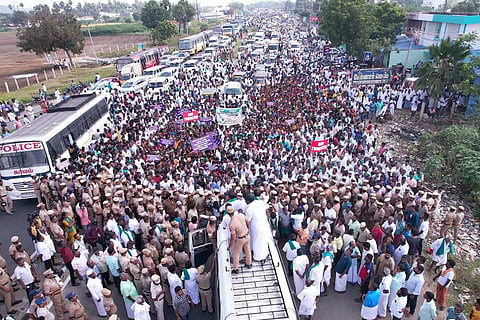 A densely packed crowd of people gathers on a road, surrounded by police officers and vehicles, including buses and cars, in a rural setting with trees and fields in the background.