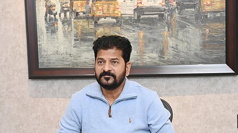 Telangana Chief Minister A Revanth Reddy in a blue full sleeved t shirt staring ahead of him. There's a painting of a road with vehicles in rain behind him. There are three water bottles and stationery on the table in front of him.