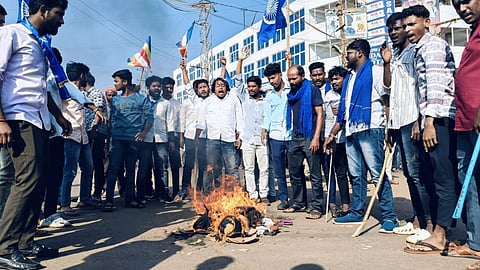 A group of people stand around a small fire on the ground during a protest on a street with buildings and signs in the background.
