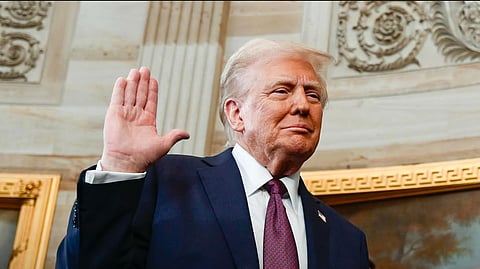 Donald D Trump with his right hand raise, facing outwards, while taking oath as US President. He is wearing a dark suit, white shirt and red tie. Paintings with gilt frame and ornate architecture can be seen behind him.
