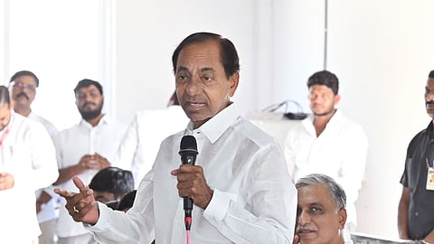 The image shows former Telangana Chief Minister KCR in a white traditional Indian outfit speaking into a microphone while gesturing with his hand. He is surrounded by several people, also dressed in white, some of whom are seated and clapping. The setting appears to be a political gathering indoors, with papers and a table visible in the foreground.