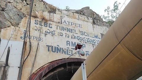 The image shows the entrance of a large tunnel with a massive pipe running through it. The entrance is labeled with text that includes "JAYPEE," "SLBC TUNNEL SCHEME," and other inscriptions in English and Telugu. There are industrial structures and equipment in the foreground, including metal scaffolding and pipes. A person is partially visible on the right side of the image. The tunnel appears to be part of an infrastructure project.