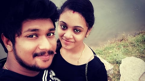 Pranay and Amrutha taking a selfie outdoors near a body of water. Pranay, who has a beard and short, wavy hair, is wearing a black graphic t-shirt and smiling slightly. Amrutha, standing beside him, has her hair tied back and is wearing a black dress with a gold chain purse, a red bindi on her forehead, and a subtle smile. The background consists of grass, rocks, and water, giving the image a natural setting.