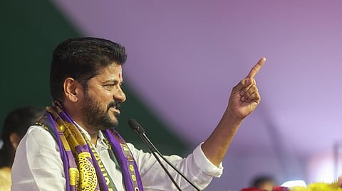 Telangana CM Revanth Reddy wearing a white shirt and a decorative purple and gold shawl is speaking into a microphone while gesturing with his right hand. He has a confident expression, a neatly trimmed beard, and a red tilak on his forehead. The background is blurred with a mix of green and purple hues, suggesting a stage or event setting.