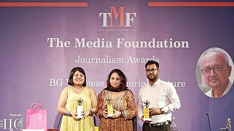 Three people stand on a stage holding awards at The Media Foundation Journalism Awards event. The backdrop displays "The Media Foundation Journalism Awards BG Verghese Memorial Lecture" along with the organization's logo and a portrait of an elderly man wearing glasses. The individuals are dressed in formal attire, smiling while posing for the photograph. A pink gift bag is visible on the side.