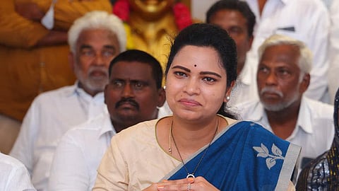 YSRCP leader Vidadala Rajini in a brown blouse and blue saree sitting in front of a bunch of mics addressing the media with her hands clasped together in front of her, with a smile on her face. There are many men in white sitting behind her in the background.