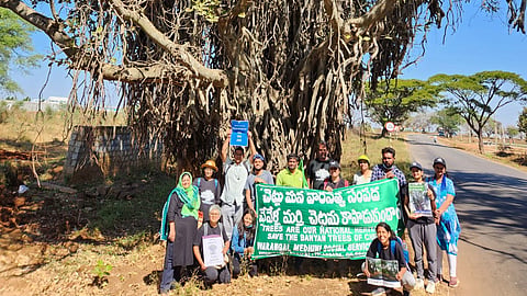 A group of people is standing in front of a large banyan tree on the side of a road, holding banners and signs advocating for the protection of banyan trees. The banners include messages in Telugu and English, with phrases like "TREES ARE OUR NATIONAL HERITAGE" and "SAVE THE BANYAN TREES OF CHEVELLA." The participants appear to be part of an environmental activism effort, and they are dressed casually, some wearing hats and sunglasses. The background features a clear blue sky and a few trees in a rural or semi-rural setting.