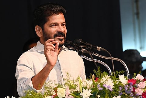 The image shows Telangana CM Revanth Reddy in a light-colored shirt speaking at a public event. He is standing behind a podium decorated with colorful flowers and is gesturing with his hand while addressing an audience. There are multiple microphones in front of him, and the background is dark with some people visible.