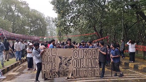 A group of people is gathered outdoors in a protest. Several individuals are holding a large cardboard sign with bold, hand-drawn lettering and an illustration of a fox. The sign reads:
"WE ARE THE FOXES"
"OUR DEN, OUR LAND, OUR RIGHTS"
The setting appears to be a university or public park, with trees, tents, and banners in the background. Many protesters are engaged in conversations, and some are looking at the sign. The atmosphere suggests activism, possibly related to land or student rights.