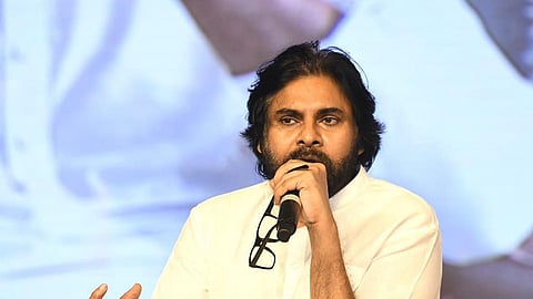 Pawan Kalyan with medium-length black hair and a beard, dressed in a white kurta, is speaking into a microphone while holding a pair of glasses. He is standing behind a clear podium, addressing an audience in a formal setting with a large screen in the background displaying his image.