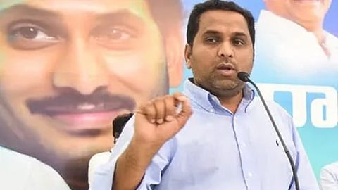 A man in a light blue shirt is speaking into a microphone at a podium. Behind him is a large poster featuring a close-up of YS Jagan's face smiling. The background suggests a political event or press conference.