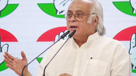 jaivam Ramesh speaking through a mic at a Congress event. He is in a white kurta. The Congress' symbol "hand" is displayed on the board behind