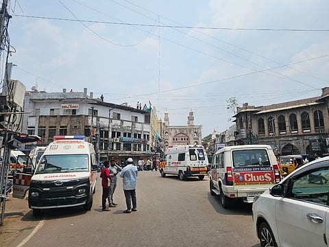 A busy street scene in Hyderabad, India, with several ambulances and a Hyderabad City Police vehicle present. People are gathered on the road, and some are seen conversing. Historic architecture lines both sides of the street, and the iconic Charminar monument is visible in the background under a partly cloudy sky. Power lines crisscross overhead.