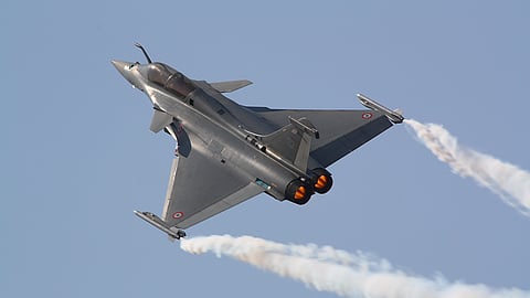 A Dassault Rafale fighter jet is seen in flight during an aerial maneuver, with both engines glowing in afterburner mode and white vapor trails streaming from its wingtips against a clear blue sky. The aircraft bears French Air Force roundels.