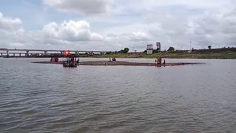 A group of people is gathered on a narrow sandbar in the middle of a river under a partly cloudy sky. A small wooden boat with red flags is anchored near the sandbar, and a concrete bridge and some structures are visible in the background on the riverbank. The scene suggests a ritual or local event taking place in a rural or semi-urban setting.