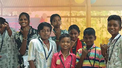 A group of smiling children and teenagers pose together, some making playful hand gestures. The background has a warm yellow and orange glow, creating a vibrant and energetic atmosphere.