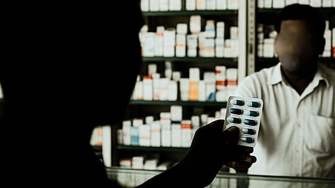 A man is holding up a strip of capsules. In front of him is a pharmacist and shelves stocked with medical drugs is seen behind the pharmacist.