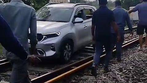 A silver SUV is on railway tracks, surrounded by several men. The scene is set in an outdoor area with trees, gravel, and railway infrastructure visible. The atmosphere is tense.