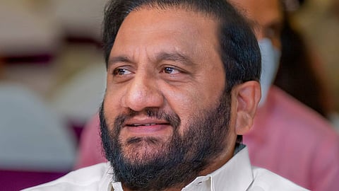 A middle-aged man with a full beard and neatly combed black hair is seen smiling slightly while looking to his left. He is wearing a white shirt and is seated indoors at what appears to be a formal or public event. The background is softly blurred, with other people and chairs faintly visible.