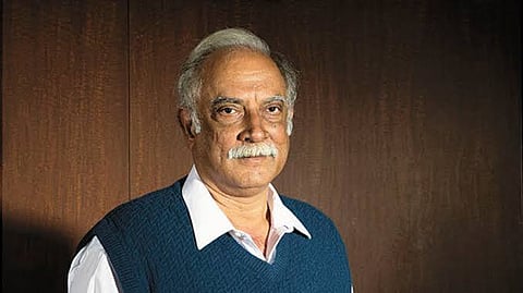 An older man with grey hair and a mustache stands against a dark wooden background. He is wearing a white shirt with a blue knitted vest over it, and he looks directly at the camera with a calm, composed expression.